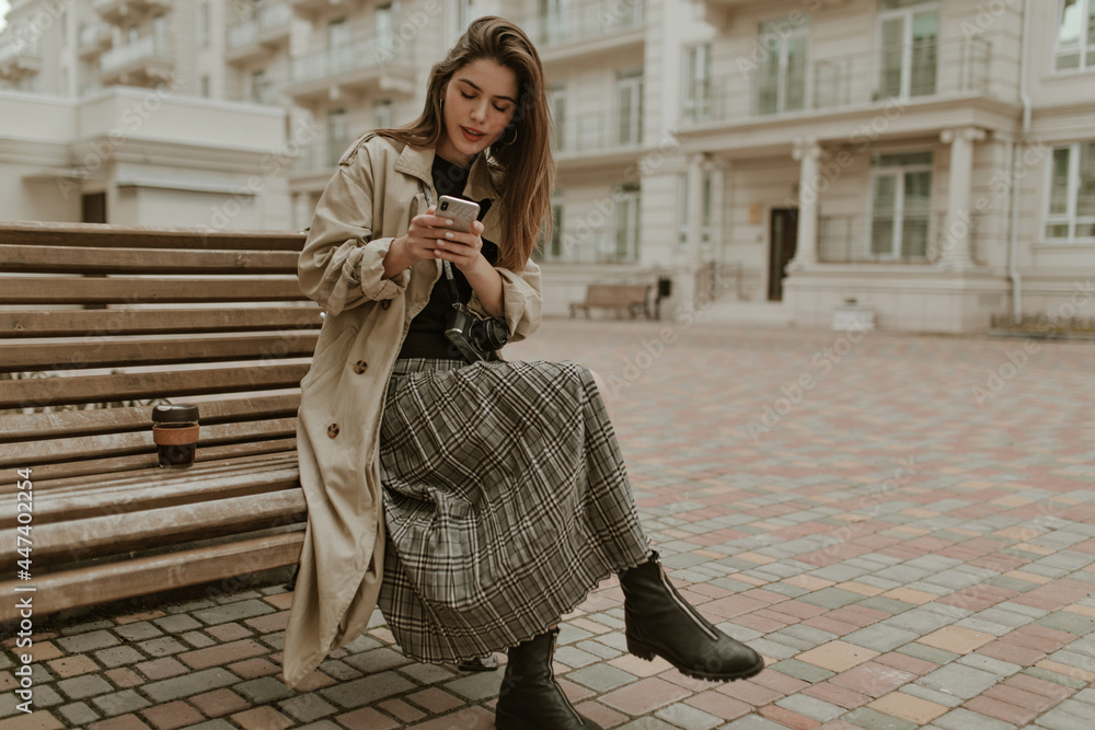 Stylish elegant woman in checkered skirt, black top and beige trench coat  sits on wooden bench outside and holds cellphone. Stock Photo | Adobe Stock