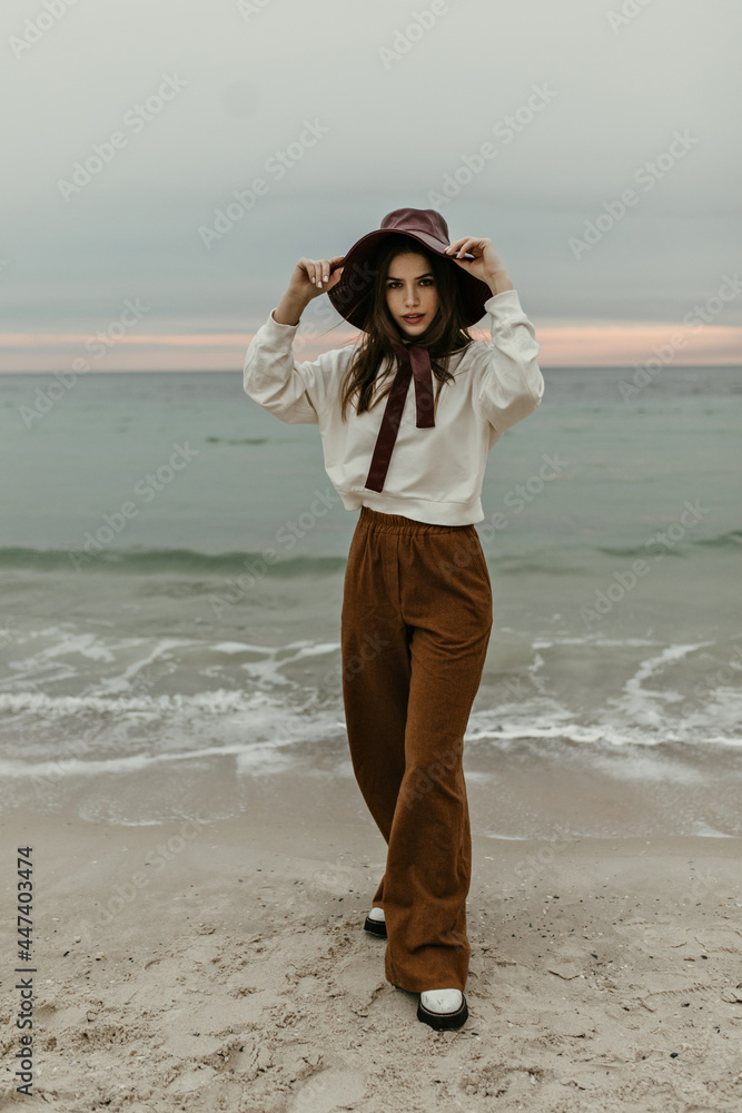 Full-length portrait of brunette woman in brown pants and beige sweater puts on wide-brimmed hat and looks into camera on sea background. Full-length portrait of brunette woman in brown pants and beige sweater puts on wide-brimmed hat and looks into camera on sea background.