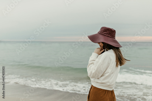 Brunette woman looks away at sea. Young attractive girl in wide-brimmed hat, brown pants and sweatshirt poses at beach.