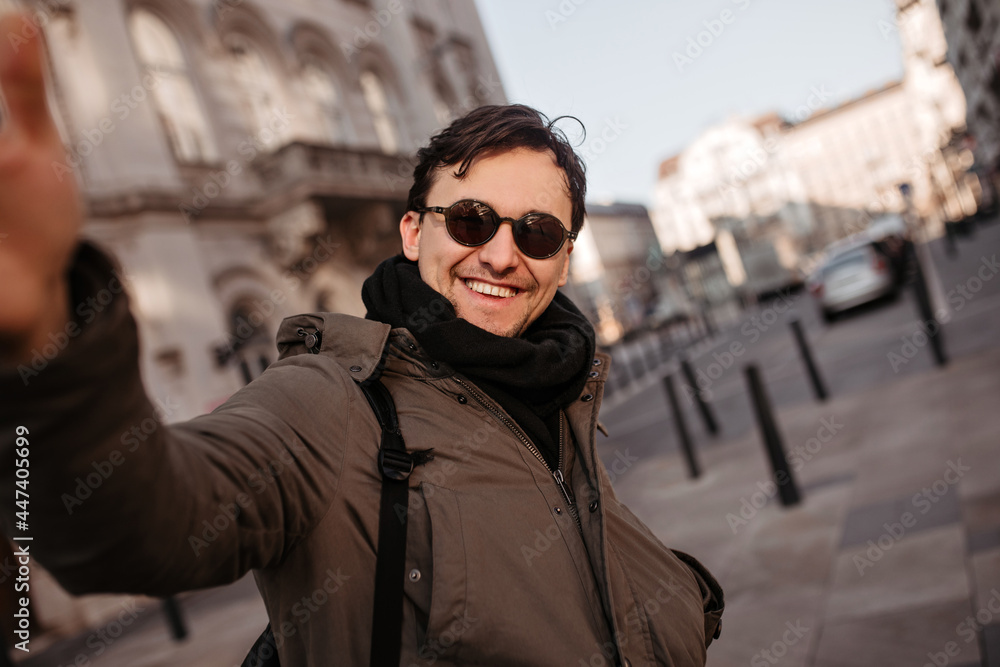 Cheerful man in sunglasses takes selfie outdoors. Brunet happy guy in khaki coat and black scarf walks around city. Cheerful man in sunglasses takes selfie outdoors. Brunet happy guy in khaki coat and black scarf walks around city.