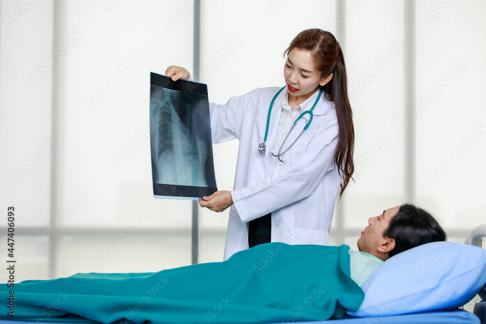 Obraz premium Young Asian female doctor demonstrating x ray scan to patient lying on bed while explaining medical treatment in modern hospital