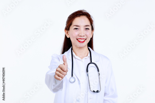 Cheerful young Asian female physician in medical uniform with stethoscope looking at camera and demonstrating thumb up gesture against white background