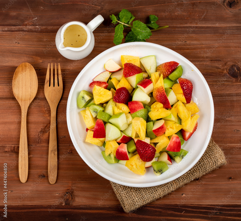 Mixed fruits salad including strawberry, kiwi, apple, and pineapple in white dish place on sackcloth on wooded table.  Utensil and oil salad dressing cup beside with mint leaf decorating