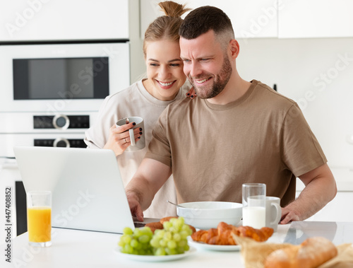 Young beautiful happy family couple reading news on laptop during morning breakfast at home