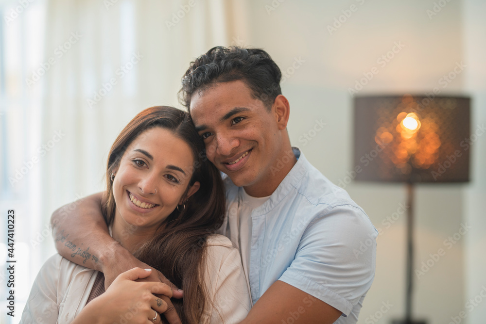 Young millennial interracial couple hug with love at home in living room - relationship with black boy and caucasian girl together standing and embracing looking each other - concept of life and house