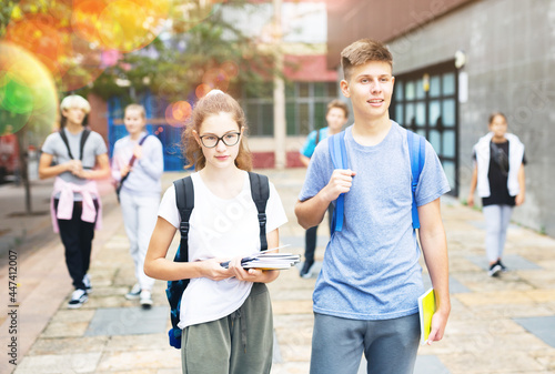 Portrait of teen female and male schoolmates with backpacks and workbooks walking to college campus on warm autumn day.