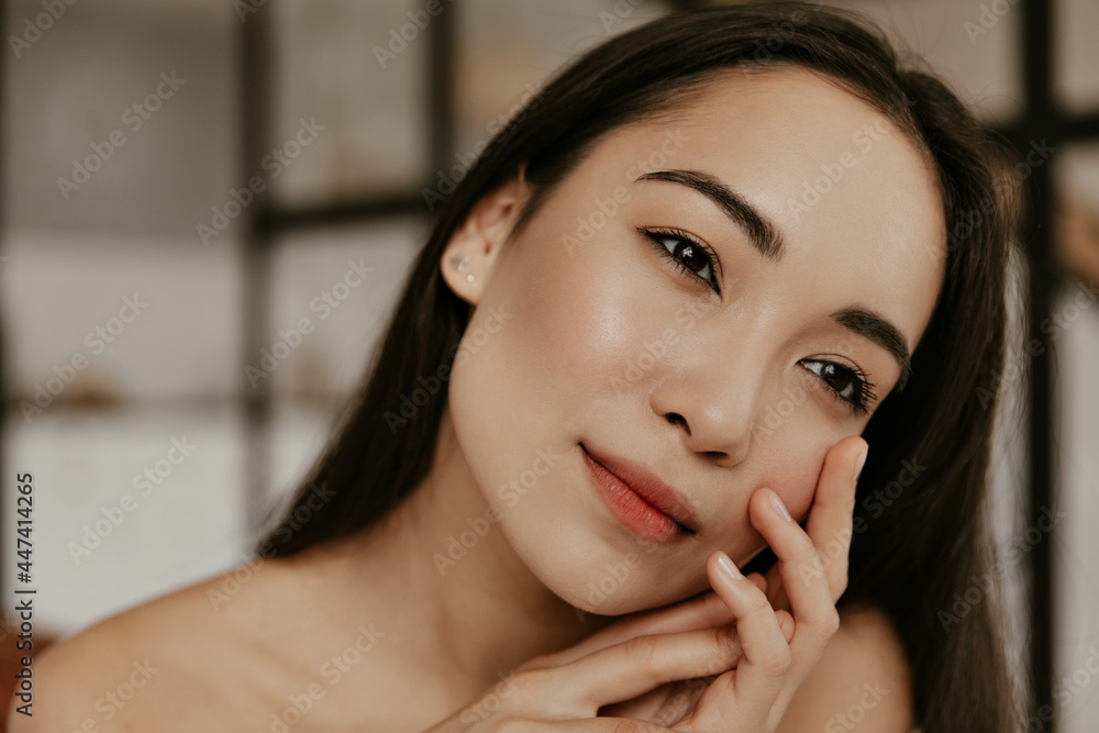 Brown-eyed brunette Asian charming woman with healthy skin looks calm and touches gently her face. Young lady poses at home. Brown-eyed brunette Asian charming woman with healthy skin looks calm and touches gently her face. Young lady poses at home.