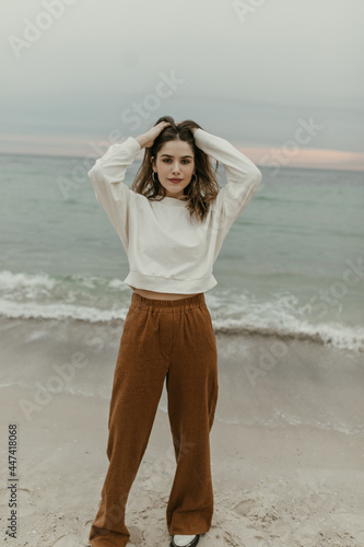 Charming girl ruffles hair and poses near sea. Attractive brunette woman in brown pants and white sweatshirt stands at beach and looks into camera.