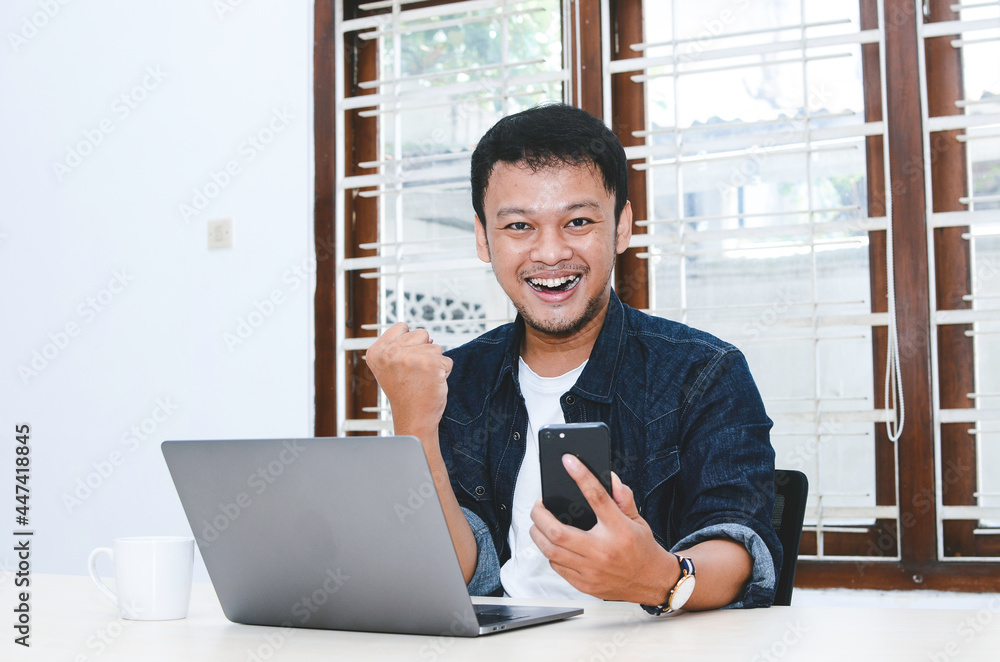 Fototapeta premium Happy excited and smiling young Asian man raising his arm up to celebrate success or achievement when working in laptop. Indonesian man wearing blue shirt.