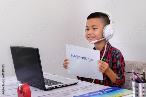 Boy sitting in front of a laptop screen, studying online at home, plugging in headphones, holding an answer sheet, showing it to the teacher