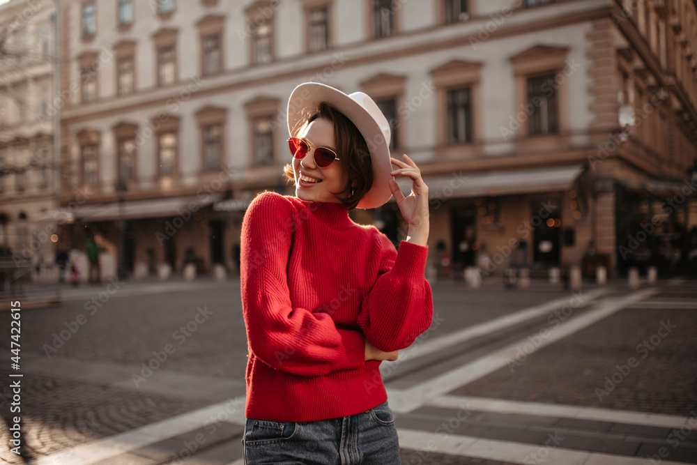 Young beautiful woman in red sunglasses smiles outside. Cheerful short-haired girl in beige hat and sweater walks outdoors.