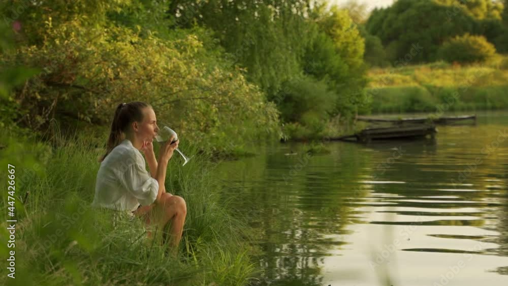 Young woman enjoying glass of wine on lake bank at sunset. Woman admiring landscape while having drink Young woman enjoying glass of wine on lake bank at sunset. Woman admiring landscape while having drink