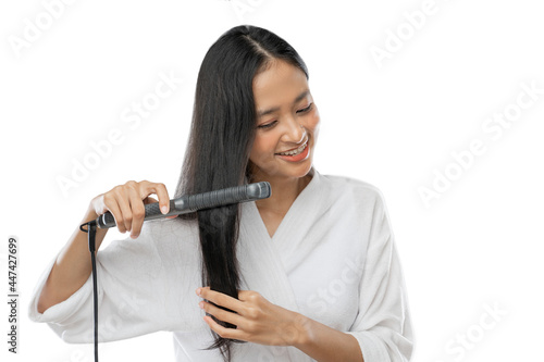 a smiling woman wearing a towel uses a hair straightener to straighten it on a light gray background