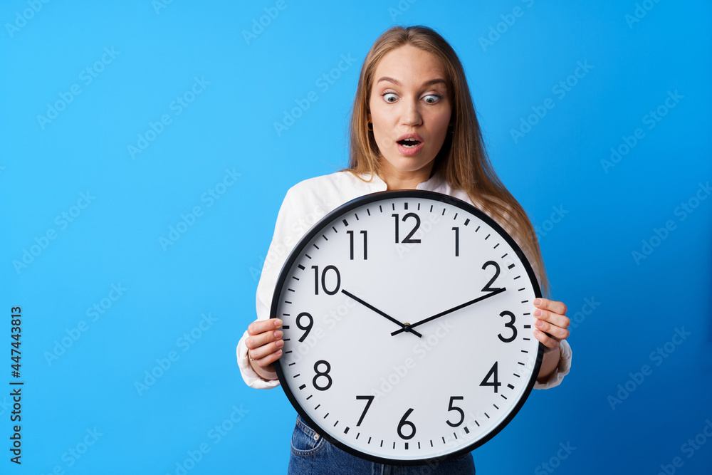 Attractive young woman holding big wall clock against blue background Attractive young woman holding big wall clock against blue background
