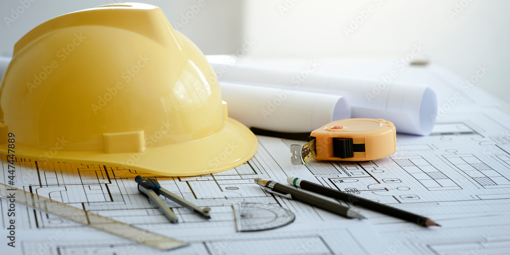 Yellow hard safety helmet hat and architectural blueprints and blueprint rolls and pencils pen, ruler, protractor, and tape measure on the table at the construction site. - obrazy, fototapety, plakaty 