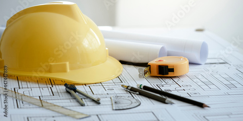 Yellow hard safety helmet hat and architectural blueprints and blueprint rolls and pencils pen, ruler, protractor, and tape measure on the table at the construction site.