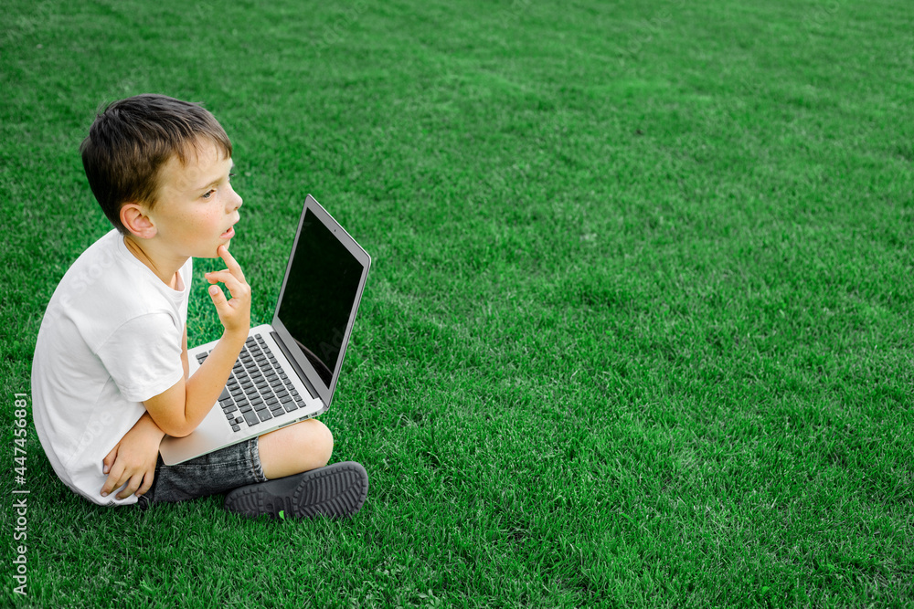 child sits on the grass and studies with a laptop online, enjoying nature child sits on the grass and studies with a laptop online, enjoying nature