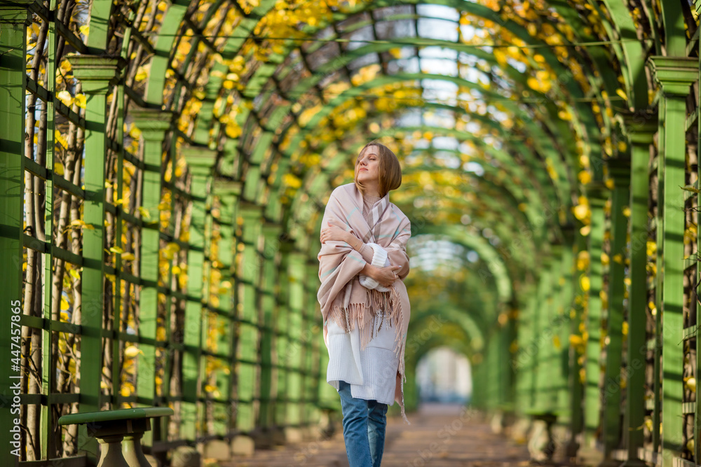 Beautiful young woman with light brown hair in a picturesque alley in autumn park. An attractive woman wrapped herself in a plaid. Fall season. Golden leaves are visible through the green fence.