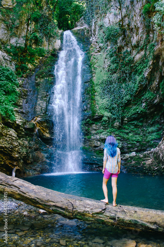 Unrecognizable young woman standing on tree trunk and looking at waterfall in summer.