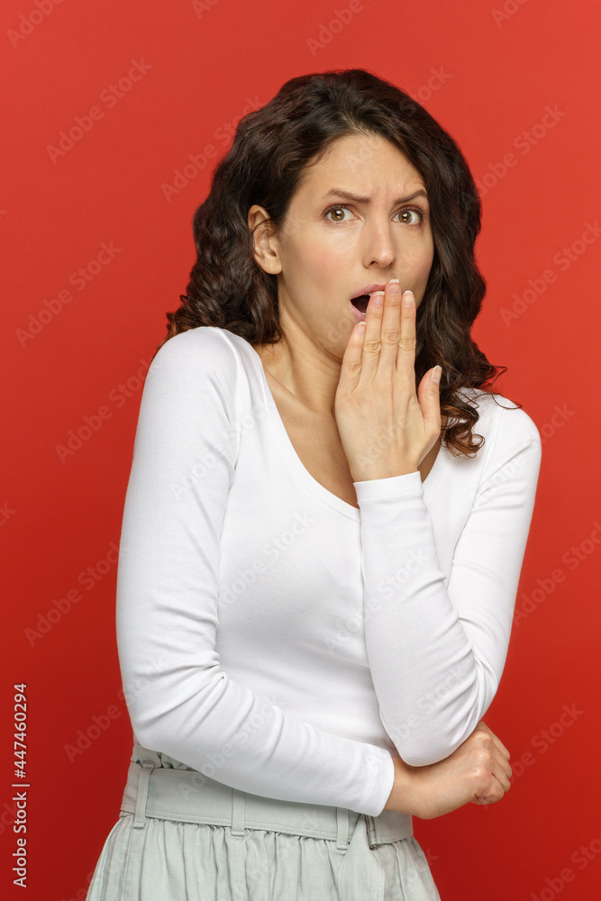 Scared shocked woman isolated on red studio wall background. Female emotional reaction to unpleasant bad news, covers mouth with her hand. Afraid frightened dissatisfied girl in 30s with amazed face Scared shocked woman isolated on red studio wall background. Female emotional reaction to unpleasant bad news, covers mouth with her hand. Afraid frightened dissatisfied girl in 30s with amazed face