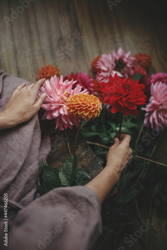 Woman in linen dress sitting on wooden rustic bench and holding dahlias flowers, view above. Rural slow life aesthetic. Autumn season in countryside. Florist arranging autumn flowers bouquet