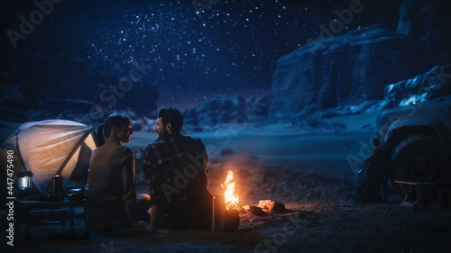 Happy Couple Nature Camping in the Canyon, Sitting Watching Campfire Together, Talking, Watching Night Sky. Two Traveling Young people On Inspirational Vacation Trip Marvel at Milky Way Stars