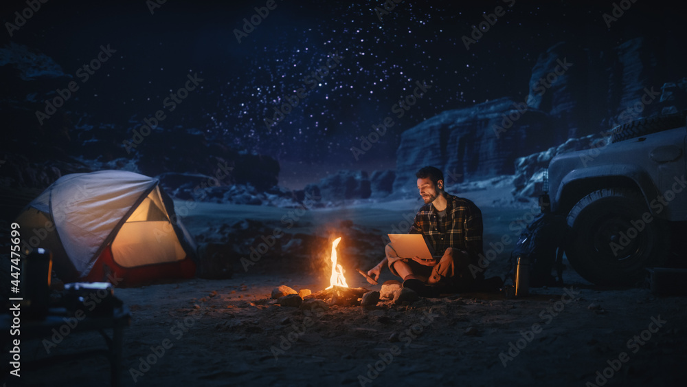 Male Traveler Sitting by Campfire Uses Laptop Computer while Tent Camping in the Canyon. Man doing Digital Remote Work, e-business, e-shopping, ecommerce through Internet while being on Vacation Trip 