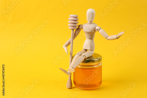 Wooden puppet holds honey spoon and sits on honey jar on yellow background