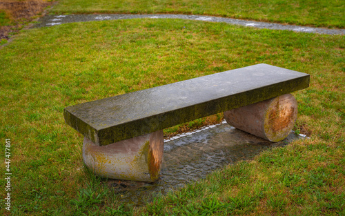 Rectangular stone bench with two round wooden tree trunks in the green meadow park.
