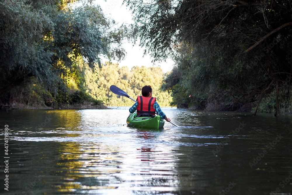 Back view on woman in green kayak paddles at wilderness river near trees at summer evening