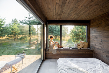 Young woman resting at beautiful country house or hotel, sitting on the window sill enjoying beautiful view on pine forest. Concept of solitude and recreation on nature