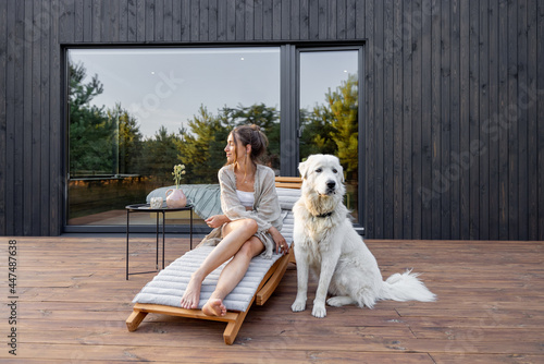 Wallpaper Mural Woman enjoys the nature while sits on sunbed on wooden terrace near the modern house with panoramic windows near pine forest whith big white dog. Concept of solitude and recreation on nature Torontodigital.ca