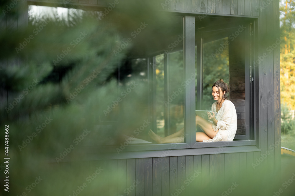Young woman resting at beautiful country house or hotel, sitting with tablet on the window sill enjoying beautiful view on pine forest. View from outside. Beautiful destinations for vacation - obrazy, fototapety, plakaty 