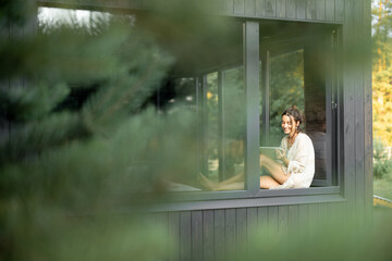 Young woman resting at beautiful country house or hotel, sitting with tablet on the window sill enjoying beautiful view on pine forest. View from outside. Beautiful destinations for vacation