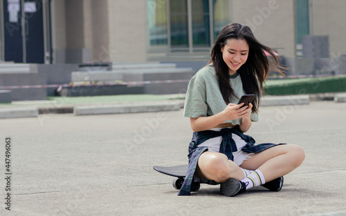 Beautiful sportive Asian female skater wearing hipster clothes, smiling with happiness, sitting on skateboard outdoor, chatting on mobile phone with copy space. Activity and Adventure Concept.