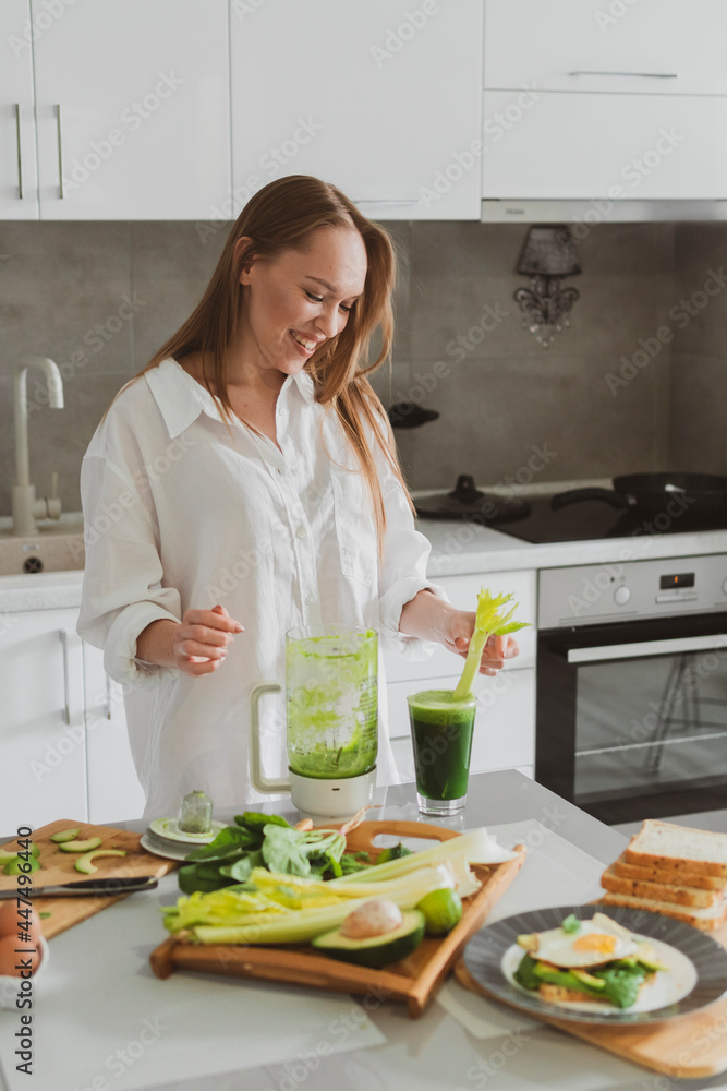 beautiful young woman preparing a healthy green smoothie in her kitchen from spinach, celery and avocado