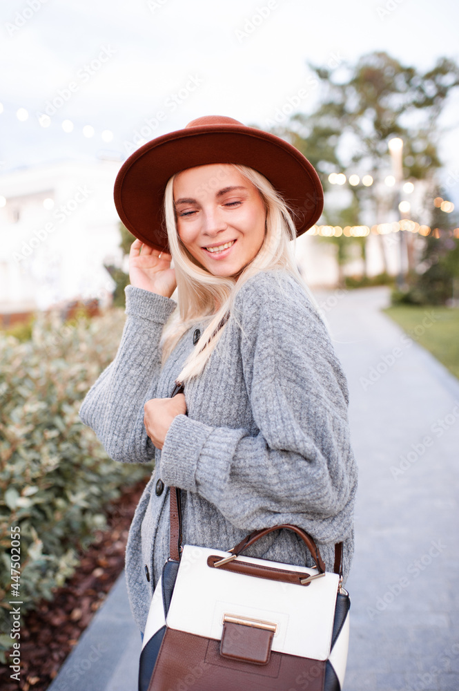 Laughing blonde cute young girl 20-24 year old wear hat, knitted woolen gray sweater and holding trendy leather bag walk in park outdoors over urban city street background. Positive emotions.