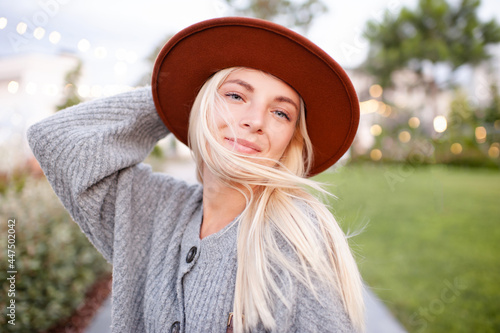 Smiling young beautiful blonde woman wear hat and knitted woolen jumper posing over city street in park outdoors close up. Happy pretty teenage girl 18-19 year old having fun outside. 20s. Happiness.