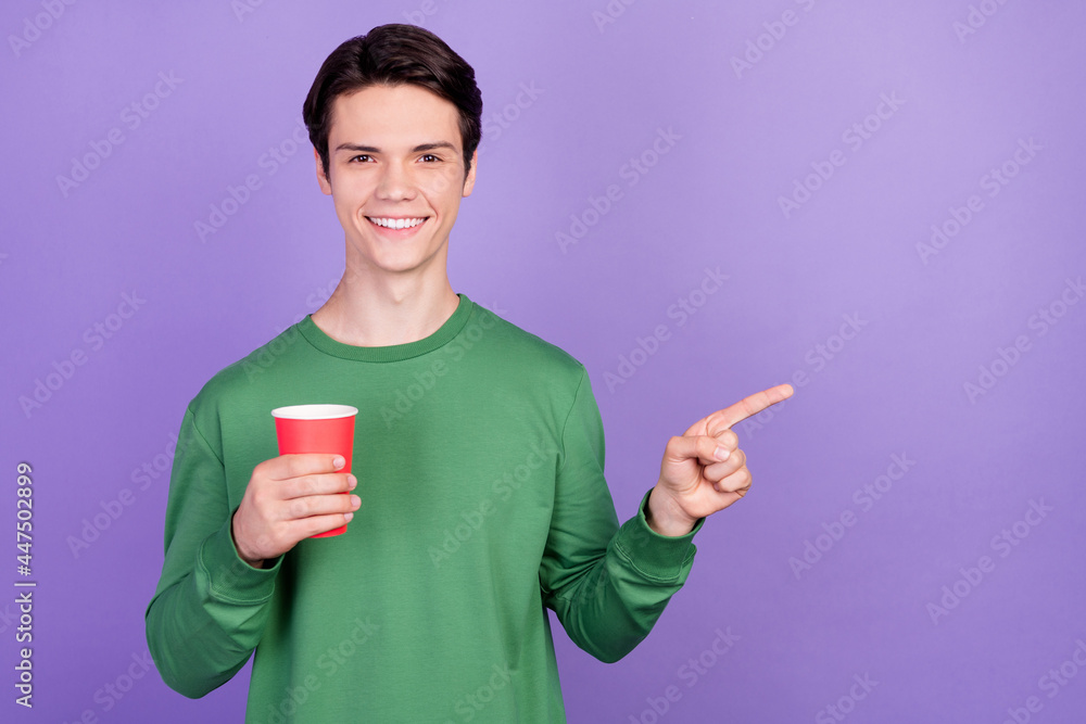 Photo of young handsome guy happy positive smile drink cup point finger empty space ad isolated over violet color background Photo of young handsome guy happy positive smile drink cup point finger empty space ad isolated over violet color background