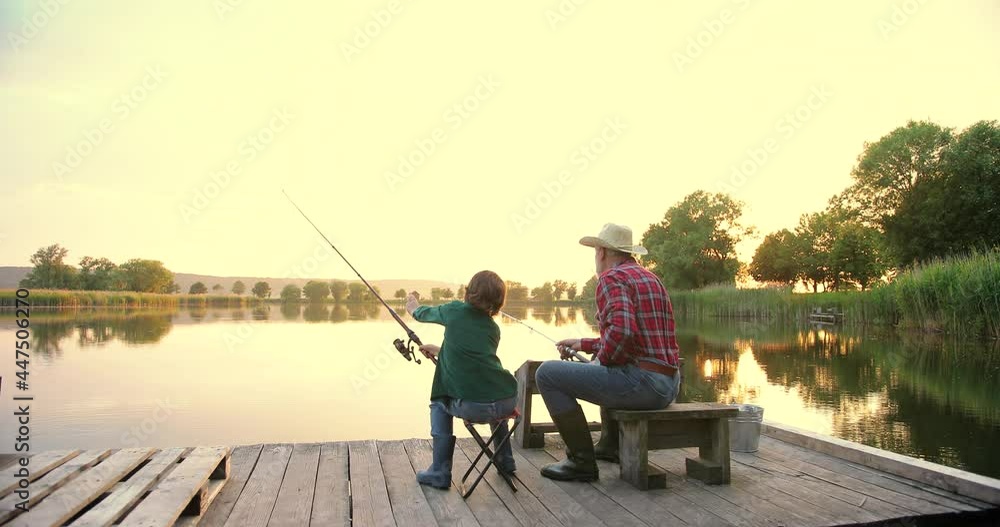 Rear view on the old grandfather and his small grandson fishing together and throwing rods in the lake while sitting on the wooden bridge or pier at the sunset Rear view on the old grandfather and his small grandson fishing together and throwing rods in the lake while sitting on the wooden bridge or pier at the sunset