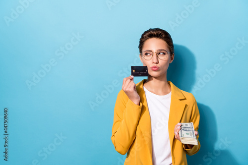 Photo of young woman pouted lips think look empty space bank card rich buy cash money isolated over blue color background