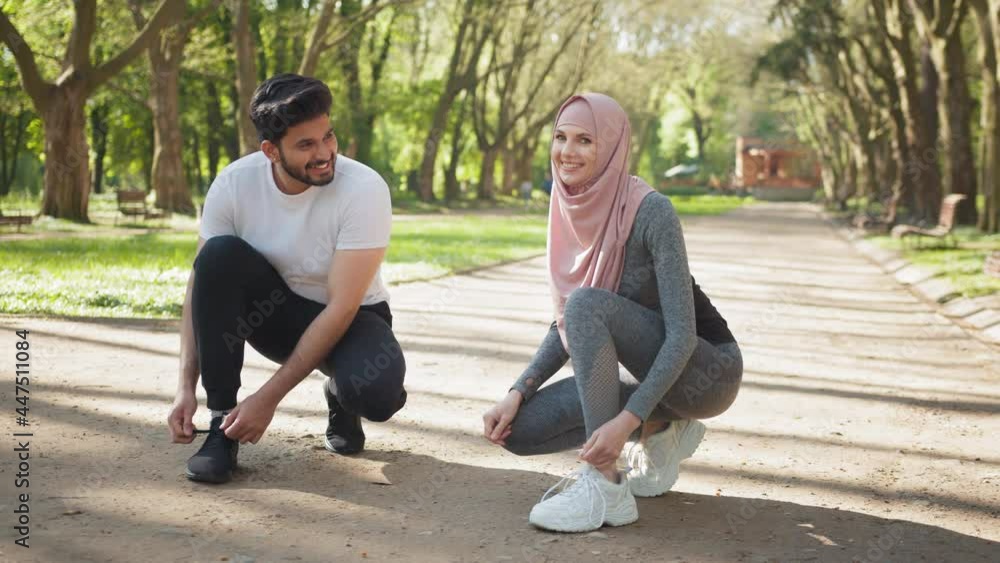 Muslim man and woman in hijab tying laces on sneakers. Happy arabian man and woman tying laces on sport sneakers, smiling and looking on each other at park. Concept of active and healthy lifestyles. Muslim man and woman in hijab tying laces on sneakers. Happy arabian man and woman tying laces on sport sneakers, smiling and looking on each other at park. Concept of active and healthy lifestyles.