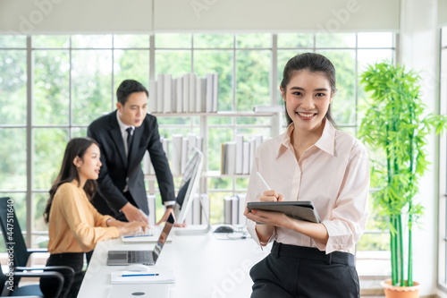 A confident young businesswoman holding tablet and looking at camera while her colleagues working in office background on business day.