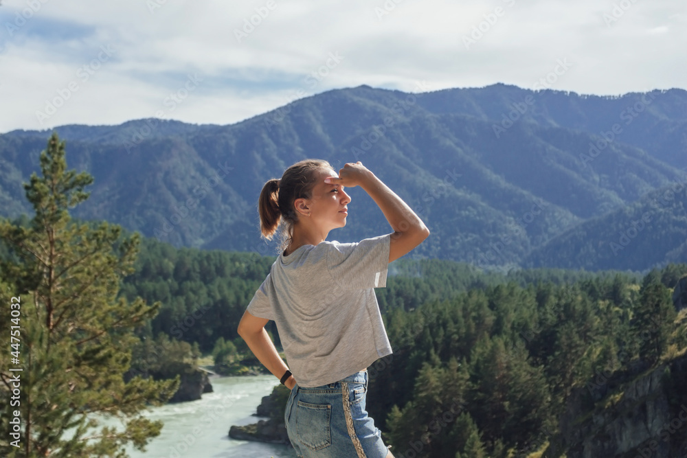 A young сaucasian slender woman looks into the distance against the background of a beautiful landscape with mountains, forests, river and a sky