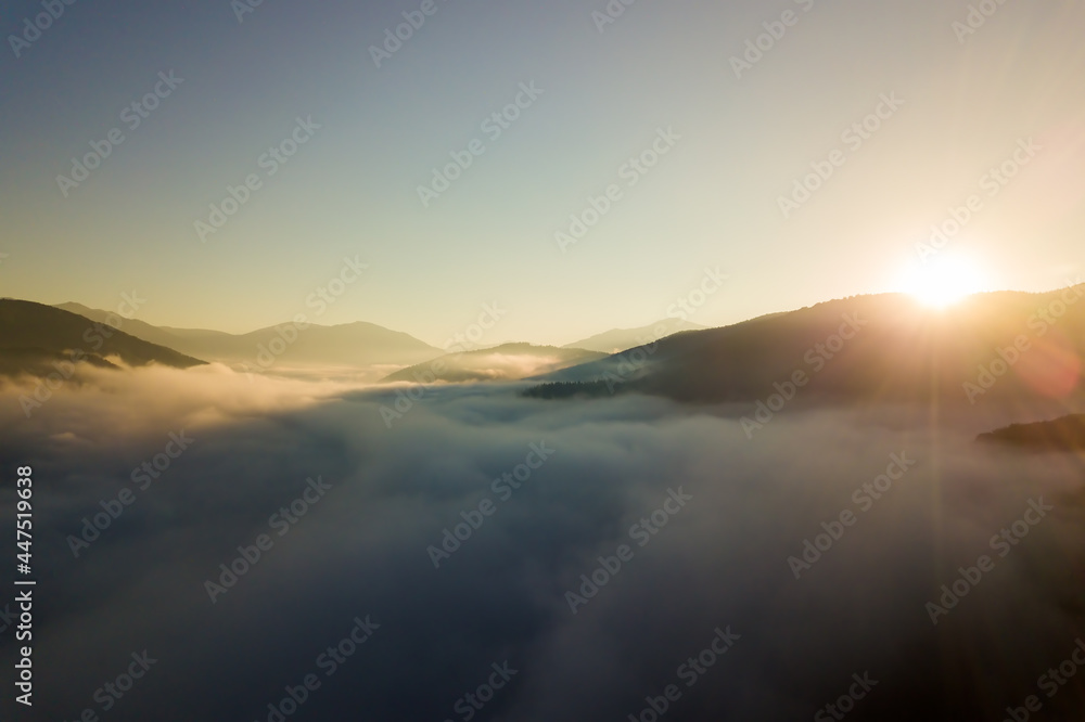 Aerial view of vibrant sunrise over white dense fog with distant dark silhouettes of mountain hills on horizon.