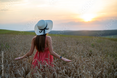 Obraz na plátne Young happy woman in red summer dress and white straw hat standing on yellow farm field with ripe golden wheat enjoying warm evening