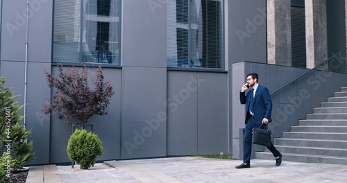 Chatting with pleasure. Full length view of happy young qualified caucasian businessman in suit is going down stairs at the street and holding mobile phone while having conversation with somebody © VAKSMANV