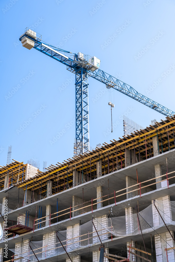 Construction crane and unfinished residential building against clear blue sky. Housing construction, apartment block with scaffolding