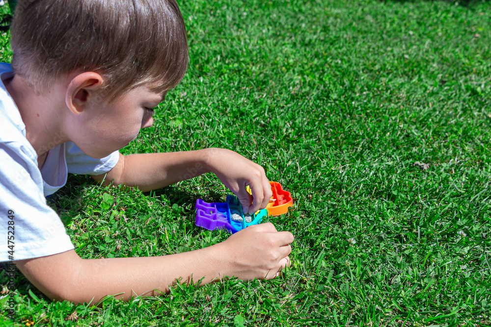 Child boy play with pop it on grass in park. New sensory developing toy in shape dinosaur with colorful rainbow silicone bubbles. Trendy fidgeting game, antistress and anxiety relief. Child boy play with pop it on grass in park. New sensory developing toy in shape dinosaur with colorful rainbow silicone bubbles. Trendy fidgeting game, antistress and anxiety relief.