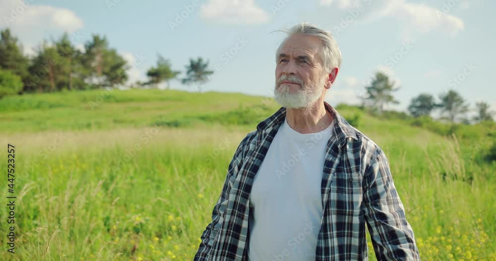Waist up portrait view of the senior smiling bearded man standing arms crossed and looking to the camera at the green field with the blue sky background during the sunny day. Real people emotions Waist up portrait view of the senior smiling bearded man standing arms crossed and looking to the camera at the green field with the blue sky background during the sunny day. Real people emotions